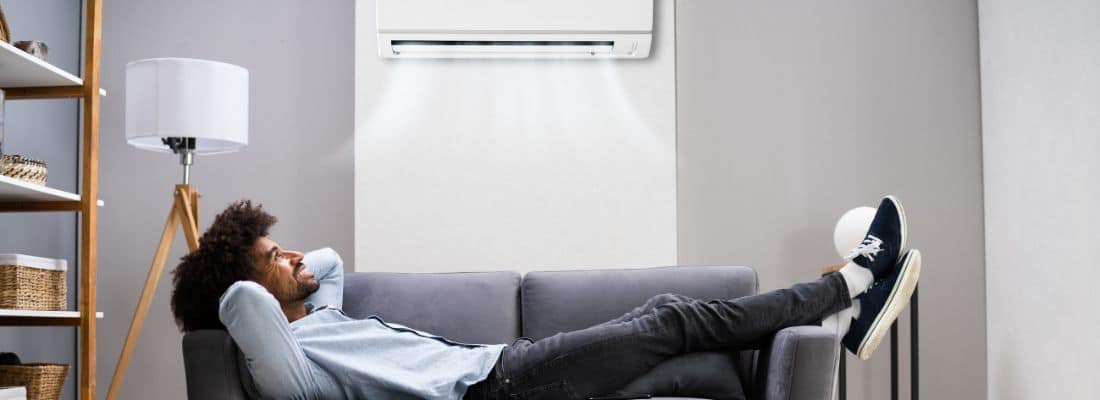 Un homme se détend sur un canapé sous un climatiseur mural, avec une lampe et une étagère en arrière-plan.